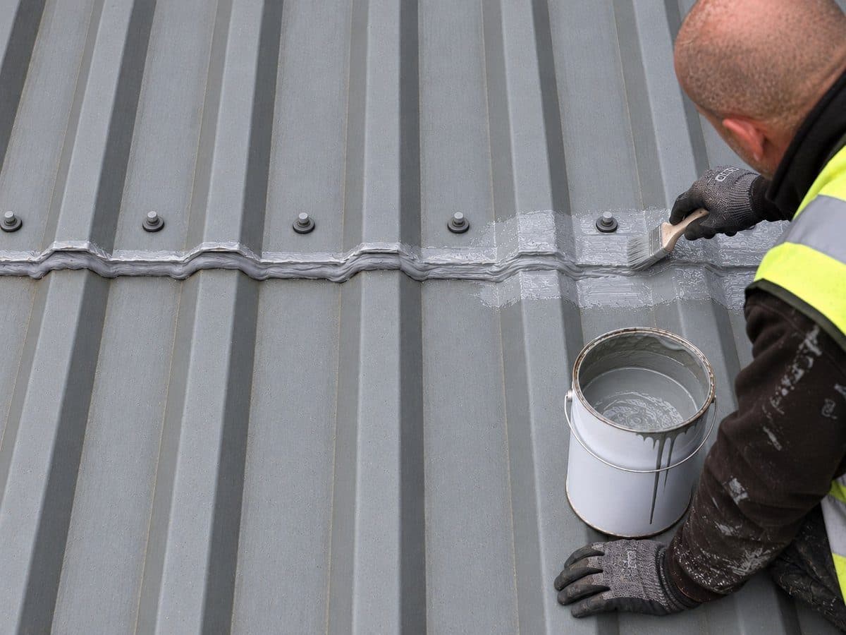 A roofing technician brushing resin over a bead of mastic along the end lap of a metal roof, with an open tin of resin beside him