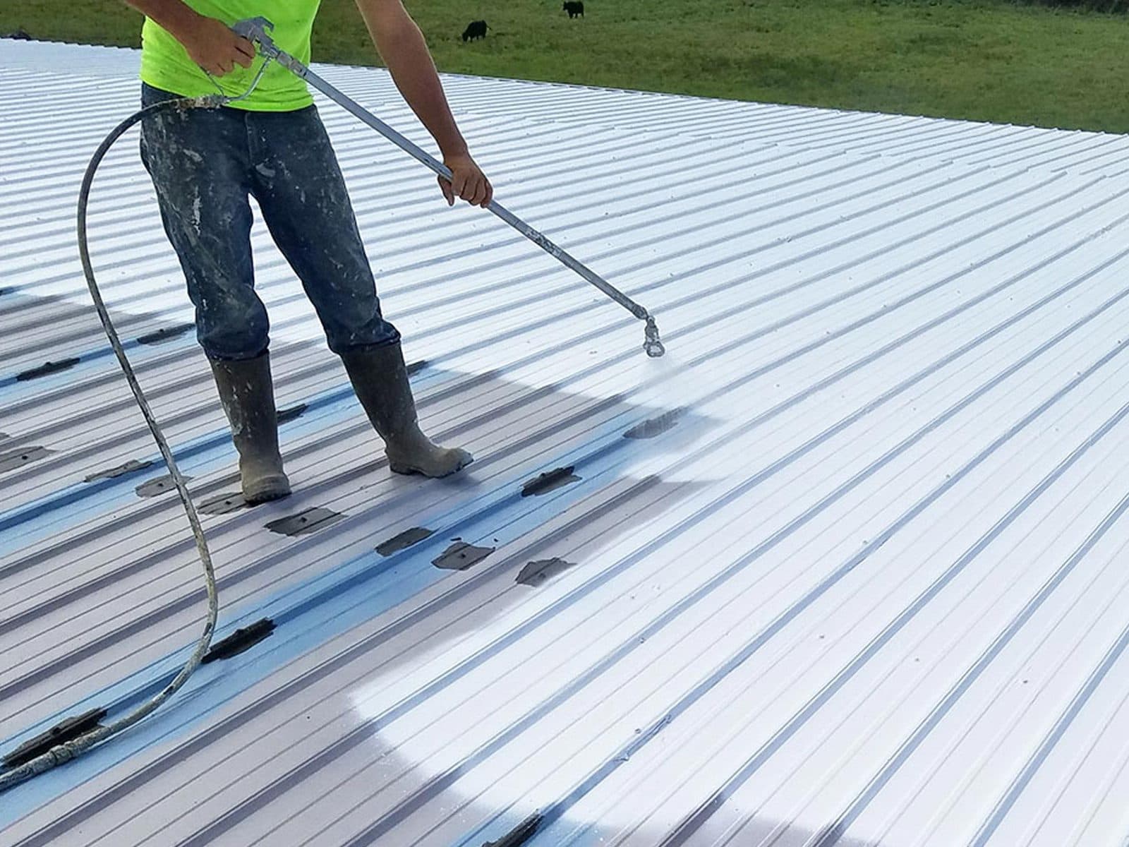A technician in hi-vis spray coating a grey standing seam roof, the sprayed area lighter behind the gun