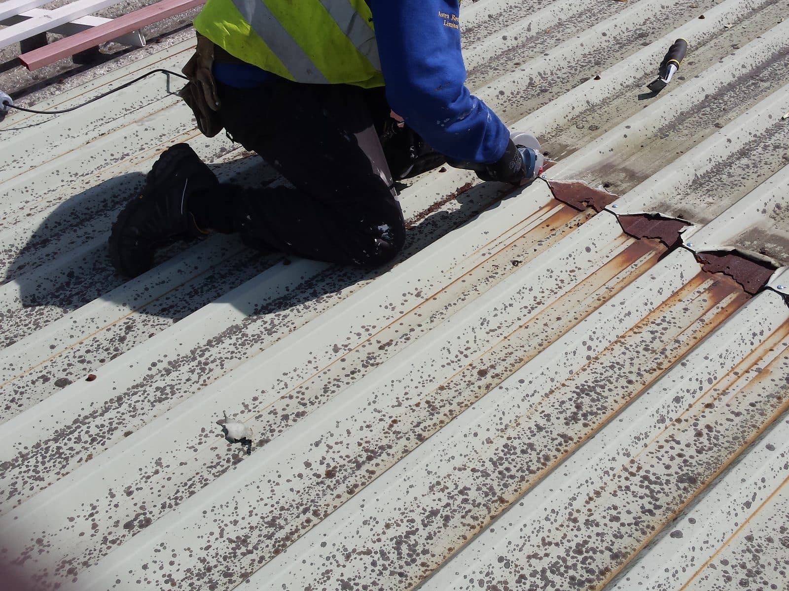 A technician kneeling at a rusted end lap on a moss-speckled profiled metal roof